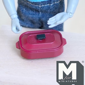 May include: A miniature burgundy casserole dish with a black handle on the lid. The dish has rounded corners and small handles on each side. The logo "M for MINIATURES" is in the lower right corner.