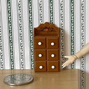 May include: A miniature brown wooden cabinet with six drawers and white knobs. The cabinet is decorated with floral accents. A silver coin rests on a wooden table. A doll is standing next to the cabinet.
