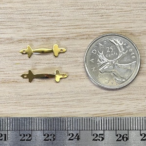 May include: Two small, gold-toned metal findings with a central bar and pointed ends, each featuring a tiny hole at both tips. These components are positioned next to a Canadian 25-cent coin and a metal ruler showing measurements in inches.