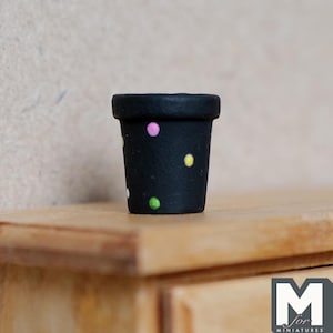 Könnte beinhalten: Ein kleiner schwarzer Topf mit bunten Punkten. Der Topf ist aus Keramik und hat eine glatte Oberfläche.