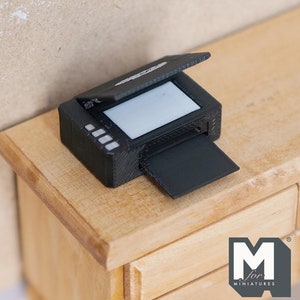 May include: A miniature black printer with a raised lid and a paper tray extending from the front. The printer sits on a light-coloured wooden surface. The printer has a white screen and a series of white buttons on the side.