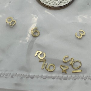 May include: A small plastic bag containing gold-coloured metal number charms. The numbers include 8, 6, 3, 7, 1, 0, and 10. A Canadian 25-cent coin is included for size reference.