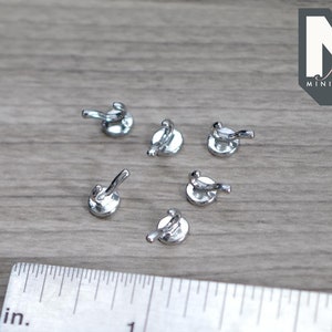May include: A collection of small, shiny silver metal miniature hooks. The hooks are arranged on a light gray wooden surface next to a ruler marked in inches and centimeters. The logo "M for MINATURES" is in the upper right corner.