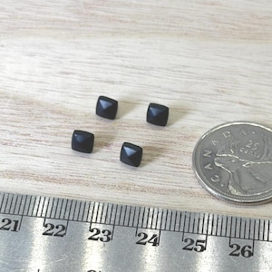 May include: Four small black square-shaped studs with a pyramid top, positioned next to a Canadian 25-cent coin and a metal ruler showing measurements in inches and centimeters to provide a clear sense of their small scale and dimensions.