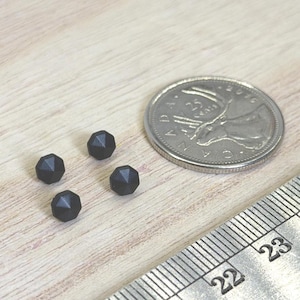 May include: Four small, matte black faceted beads arranged in a cluster next to a Canadian quarter coin and a metal ruler showing measurements in centimeters, providing a clear visual scale for the size of these individual craft components.