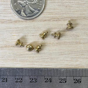 May include: Five small gold-toned metal pin backs arranged near a Canadian 25-cent coin and a metal ruler showing measurements in centimeters and inches, providing a clear scale for these tiny jewelry components used for securing pins.