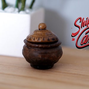 May include: A small, brown, ceramic pot with a lid. The pot is sitting on a wooden surface. The text "Ship from Canada" is visible in the background.