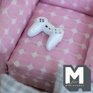 May include: A white plastic miniature video game controller on a pink and white fabric with white polka dots. The fabric is part of a miniature armchair. The image includes the text "M for Miniatures".