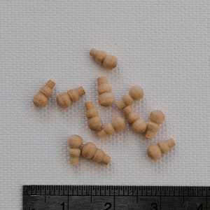 May include: A collection of small, unfinished wooden dollhouse furniture pieces, resembling miniature chairs or stools. A ruler with centimetre markings is placed next to the pieces for scale. The pieces are light brown in colour.