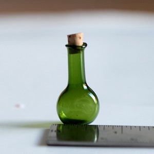 Puede incluir: Una pequeña botella de vidrio verde con un tapón de corcho. La botella mide aproximadamente 5 cm de alto y está sobre una regla.