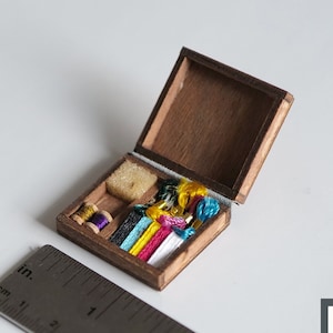 May include: A miniature wooden sewing box with a hinged lid, open to reveal spools of colorful thread, a small piece of fabric, and a small piece of white sponge. The box is about 2 inches wide.