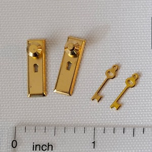 Könnte beinhalten: Miniatur-Türbeschlagsatz in Gold, bestehend aus zwei Türschildern mit Knöpfen und Schlüssellöchern sowie zwei kleinen Bartschlüsseln. Ein Lineal zeigt die Maße in Zoll. Das Logo "M for Miniatures" befindet sich oben rechts.