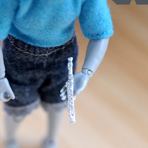 Può includere: Un flauto argentato in miniatura tenuto da una bambola grigia articolata che indossa una maglietta blu e pantaloncini di jeans. La bambola è posizionata davanti a una superficie di legno chiaro. Il logo "M for MINIATURES" è visibile in alto a destra.