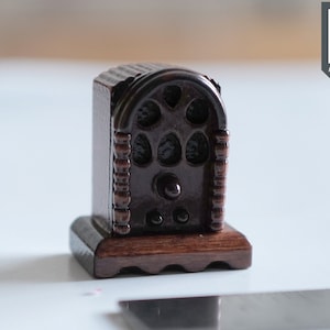 May include: A miniature wooden replica of a vintage radio with a dark brown finish and intricate details. The radio has a rounded top with a speaker grille and a dial with knobs. The base is a dark brown wood with a decorative edge.