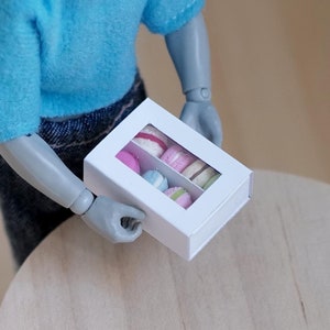 May include: A white box with a clear window, containing miniature macarons in pink, blue, and white. The box is being held by a person wearing a blue shirt and jeans.