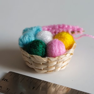 Op de afbeelding: Een klein rieten mandje gevuld met kleurrijke garenbollen, waaronder roze, geel, wit, blauw en groen. Het mandje is ongeveer 5 cm in diameter.