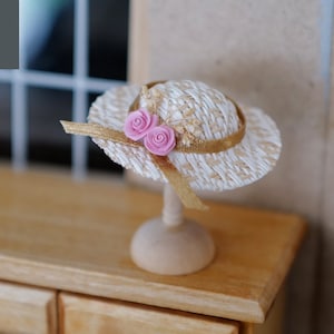 May include: A miniature straw hat with a white and brown woven design and a gold ribbon. The hat has two pink fabric flowers on the front. The hat is displayed on a wooden stand.