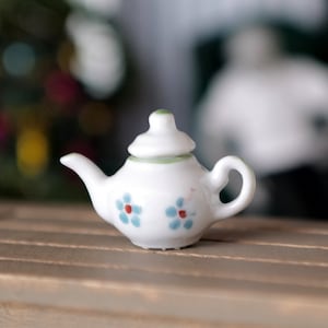 May include: A miniature white teapot with a green lid and blue and red floral design. The teapot is sitting on a wooden surface.