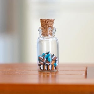 May include: A small glass bottle with a cork stopper filled with colourful miniature tiles. The bottle is sitting on a wooden table. M for Miniatures is printed on a grey square in the top right corner of the image.