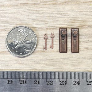 May include: Two miniature copper-colored keys and two rectangular door plates with knobs, displayed next to a Canadian 25-cent coin and a metal ruler showing measurements in centimeters and inches for scale reference.