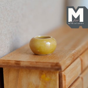 Könnte beinhalten: Ein kleiner, gelber Keramiktopf steht auf einer Holzoberfläche. Der Topf ist glasiert und hat einen leichten Glanz. Die Oberfläche ist aus Holz und hat eine natürliche Oberfläche.