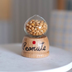 May include: A miniature glass jar filled with small, light brown peanuts, sitting on a wooden base with the word "Peanut" written on it. The jar and base are on a white table.