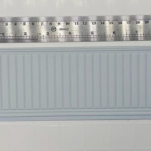 Könnte beinhalten: Ein hellblaues, rechteckiges Miniatur-Wandsegment mit vertikalen Rillen, neben einem Lineal mit Zollangaben und einer Münze. Das Lineal misst etwa 23 cm in der Länge. Das Wandsegment befindet sich auf einer weißen Fliesenoberfläche.