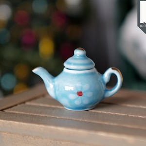 May include: A miniature blue teapot with a white flower and red centre design. The teapot has a gold rim and is sitting on a wooden table.