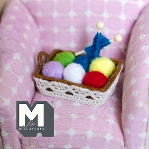Op de afbeelding: Een miniatuurmandje gevuld met kleurrijke bollen garen in groen, paars, wit, geel en rood, samen met blauwe breinaalden. Het mandje staat op een roze fauteuil met witte stippen. Het logo "M for MINIATURES" is zichtbaar.