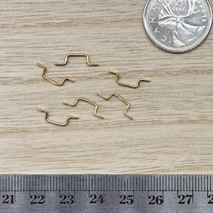 May include: Five small, gold-toned metal wire staples with a stepped, zig-zag shape. A Canadian 25-cent coin is positioned above the staples for scale, and a metal ruler showing both inches and centimeters is placed along the bottom edge.