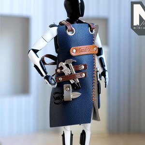 May include: A miniature artisan's apron in navy blue leather, adorned with miniature tools like a hammer, scissors, and a knife. The apron is accented with brown leather straps and the words "Hand Made". The figure is white and black.
