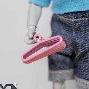 May include: A set of five pink plastic hangers, stacked together. The hangers are designed for miniature dolls or action figures.