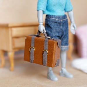 May include: A miniature brown suitcase with gray straps and gold-colored buckles is held by a doll figure. The doll is wearing a light blue sweater and denim shorts. The logo "M for MINIATURES" is in the upper left corner.