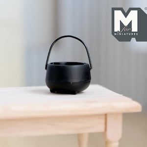 May include: A black miniature cauldron with a black handle. The cauldron is sitting on a light wood table.
