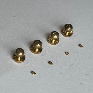 May include: Four gold-coloured shirt studs with a round top and a flat base, plus several small, oval-shaped fasteners. The studs are arranged diagonally, suggesting a set for formal wear or a tuxedo.