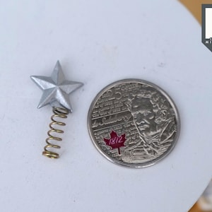 May include: A silver star-shaped tree topper with a gold spring coil. The topper is shown next to a Canadian 25-cent coin featuring a portrait of Sir John A. Macdonald and the text "1812".