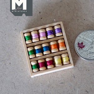 Può includere: Un set di mini bobine di filo da cucito in legno con bobine di filo colorate. Le bobine sono disposte in una scatola di legno. Una moneta d'argento con un ritratto e testo è accanto alle bobine di filo. Il logo "M for miniatures" è in alto a sinistra.