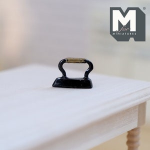 May include: A miniature black iron with a gold handle. The iron is sitting on a white wooden table.