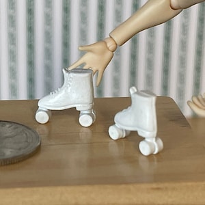 May include: A pair of white miniature roller skates with black wheels. The skates are on a wooden surface next to a silver coin.