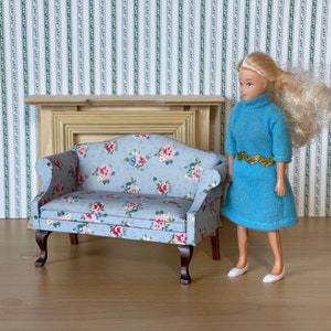 Peut inclure: Un canapé miniature bleu à motifs floraux avec des pieds en bois. Le canapé est devant une cheminée de maison de poupée et une poupée vêtue d'une robe bleue et d'une ceinture dorée se tient à côté.
