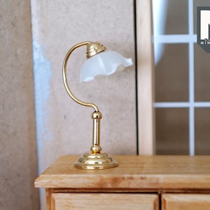 Può includere: Una lampada da tavolo dorata con un paralume in vetro bianco. La lampada è appoggiata su una cassettiera in legno con cassetti.