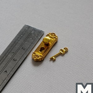 Op de afbeelding: Vergulde miniatuur deurknop en sleutelset met sierlijke details. De knop heeft een ronde handgreep en een sleutelgat. De sleutel is klein en heeft een eenvoudig ontwerp. De set is ongeveer 2,5 cm lang.