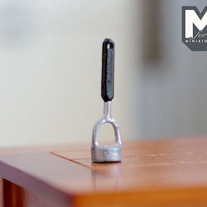 May include: A miniature silver potato masher with a black plastic handle. The masher is sitting on a wooden table.