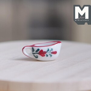 May include: A miniature white ceramic creamer with a red rim and handle. The creamer is decorated with red floral and green leaf designs. The creamer is on a light wood surface. The logo "M for MINATURES" is in the upper right corner.