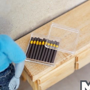 Pode incluir: Uma caixa de cigarros em miniatura com uma tampa de plástico transparente. A caixa está cheia de cigarros marrons e dourados. A caixa está sobre uma superfície de madeira.