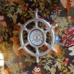 May include: A silver clock with a ship's wheel design. The clock face has Roman numerals and a compass rose. The background is a dark floral pattern with owls, deer, and birds.
