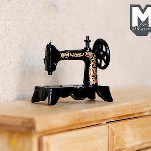 Op de afbeelding: Een miniatuur zwarte naaimachine met gouden accenten en een decoratief ontwerp. De machine staat op een houten oppervlak met laden.