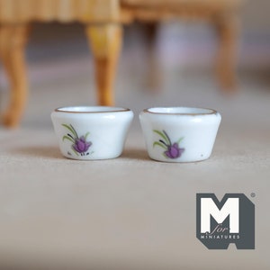 May include: Two small white ceramic bowls with a gold rim and a purple flower design. The bowls are on a light gray surface.