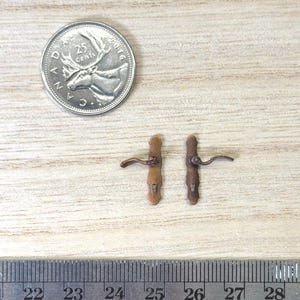 May include: Two miniature bronze-colored door handles with backplates, positioned next to a US quarter coin and a metal ruler showing measurements in inches, indicating the handles are approximately 0.75 inches in total length.