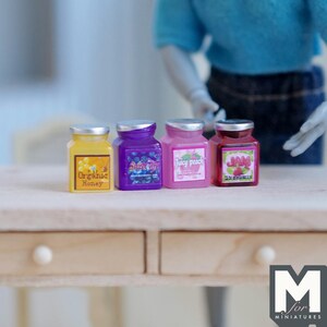 May include: Miniature jars of honey, blueberry, peach, and strawberry jam sit on a wooden table. The jars are yellow, purple, pink, and red, with silver lids and labels. The text "Organic Honey," "Juicy Peach," and "Strawberry Jam" are visible.